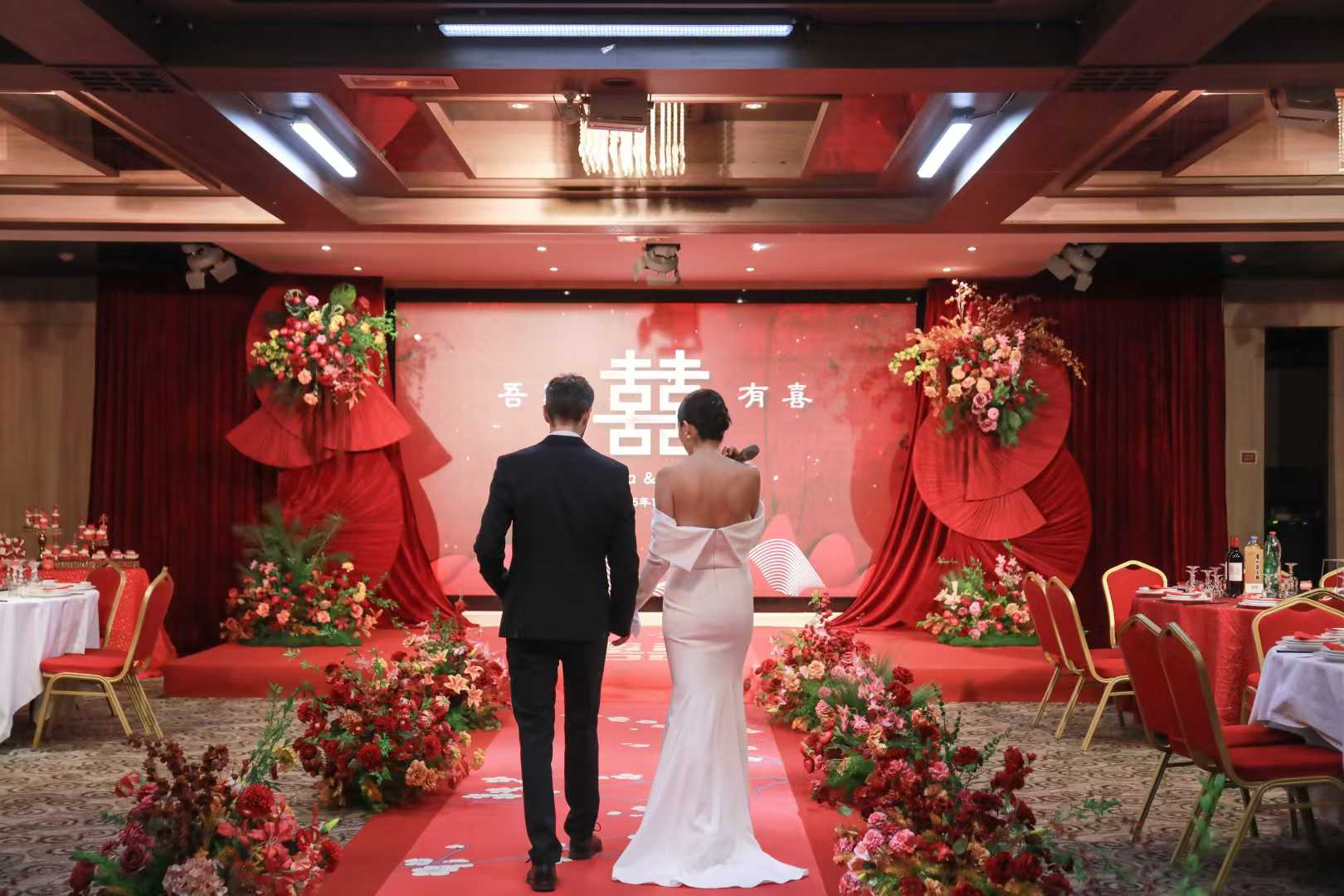 Bride and groom walking down a red carpet aisle toward a red double-happiness ceremony backdrop